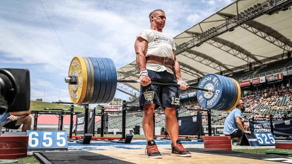 Man performing a 655-pound deadlift at a CrossFit competition in an outdoor stadium, using a Rogue barbell with bumper plates.