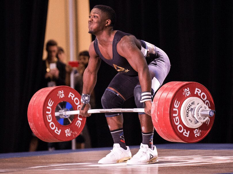 Muscular Black man in a black and white singlet performing a snatch with a barbell on a competition stage.